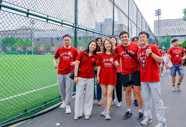 Students in Peking University shirts pose in front of a green sports field.