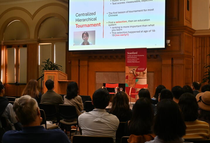 A full audience seated in front of a screen with a virtual speaker on it and presentation loaded.
