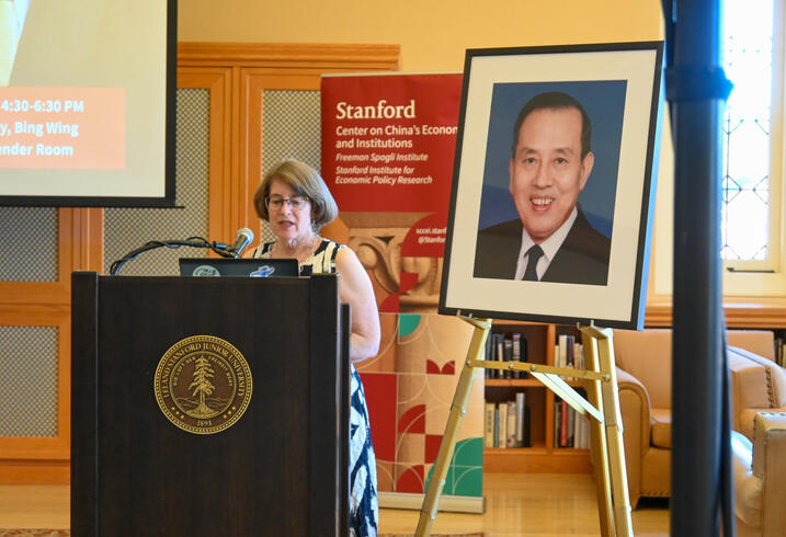 Julie Sweetkind-Singer speaks at the front of a room behind a podium and a large portrait of a man.
