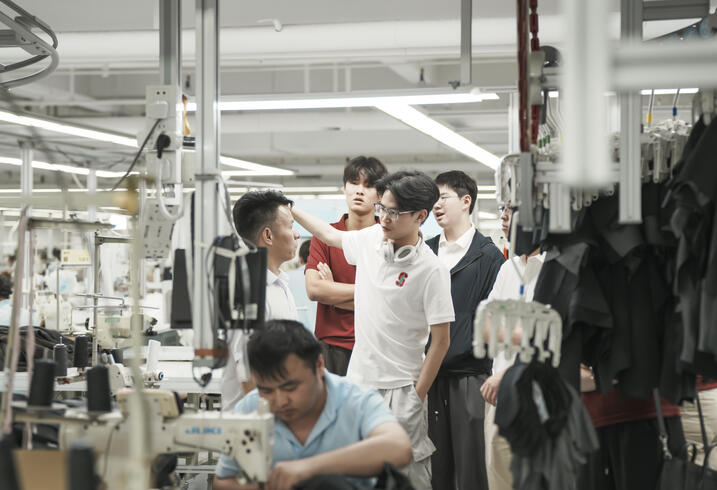 Students participate in a tour on the Yaying factory tour. A man sews in the forefront on a sewing machine.