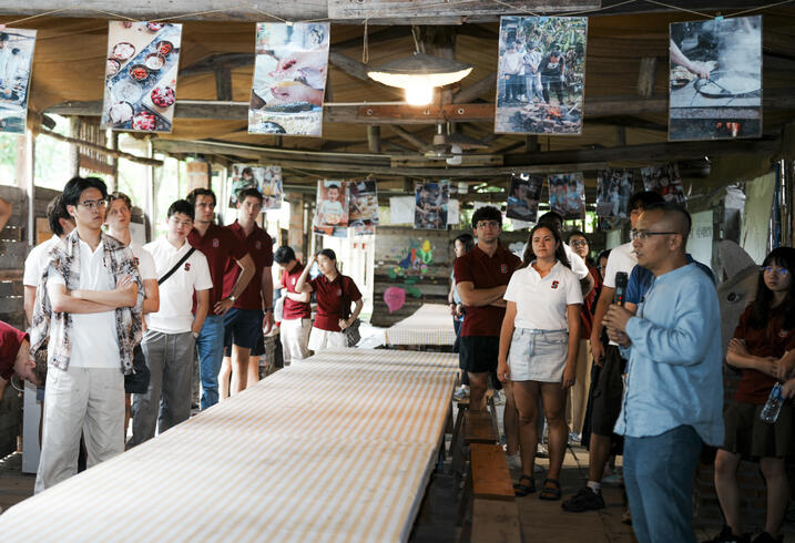 Students stand in a crowded room with someone speaking on a microphone.