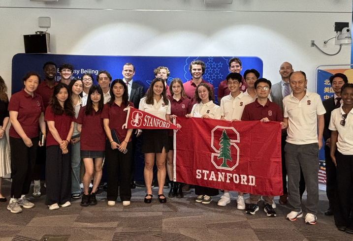A group photo of students an faculty standing with a Stanford flag and an embassy banner.