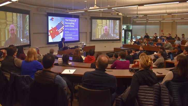 Jean Oi at a lectern introducing the panelists of a session about U.S.-China decoupling in front of a  room packed with audience members. 