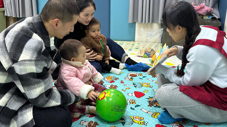 Parents hold two babies while caretaker reads to them. 
