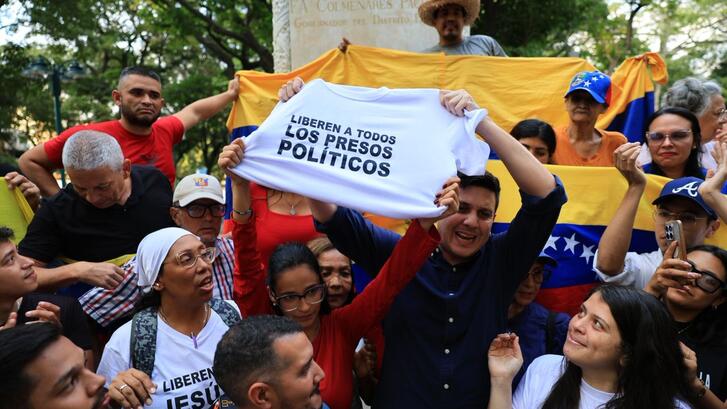 Jesús Armas stands in a crowd in front of a Venezuelan flag following his release from prison, holding a t-shirt that reads "Release all political prisoners" in Spanish.