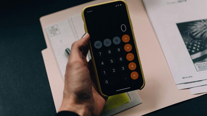 A hand holding an iPhone in a yellow case displaying the iOS Calculator app with a black screen, orange operator buttons, and gray function buttons, resting over papers and documents on a dark desk.