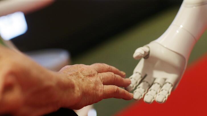 Elderly woman's hand touching a robot's hand. Photo from a summit on AI's potential for empowering humanity.