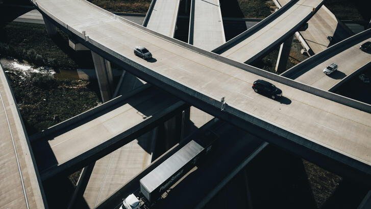 Aerial view of a multi-level highway interchange with sweeping curved overpasses crossing above one another. A few cars and a white semi-truck travel on the mostly empty concrete roads, while patches of vegetation and water are visible below the elevated ramps.