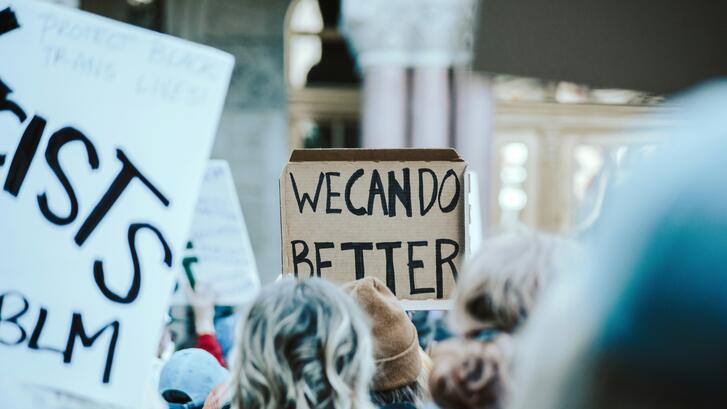 A crowd of people gathers at a protest, holding handmade signs. A cardboard sign in the center reads “WE CAN DO BETTER.”