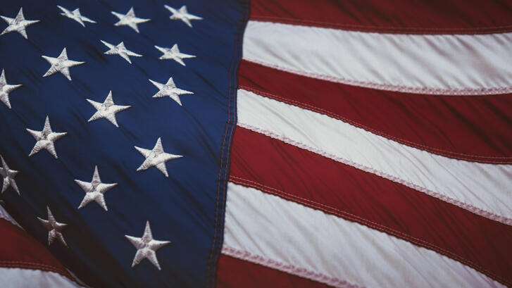 Close-up photo of a rippling American flag, with embroidered white stars on a blue field on the left and red-and-white stripes waving on the right.