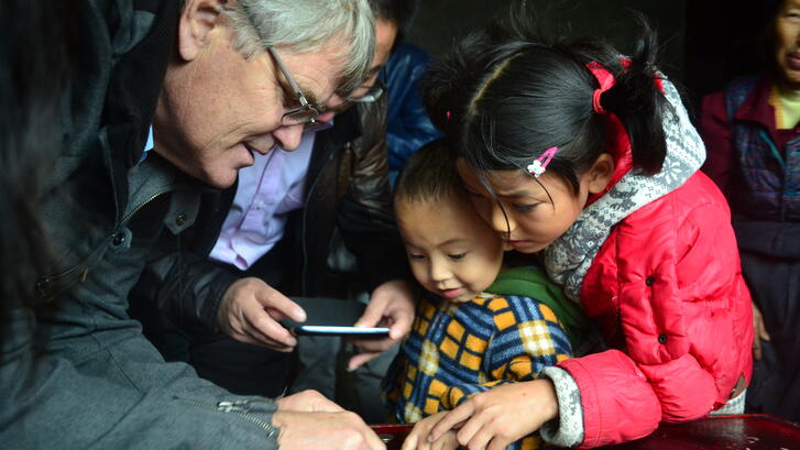 Scott Rozelle huddles together with two young children around a table looking at a phone screen and pointing to something on the table.