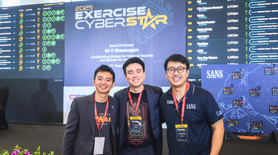 Photo of MIP Student RJ Teoh posing with two others in front of a banner that reads 2025 Exercise Cyber Star