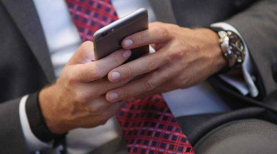 Close-up of a person in a suit and red patterned tie using a smartphone, with a wristwatch visible on their left wrist.