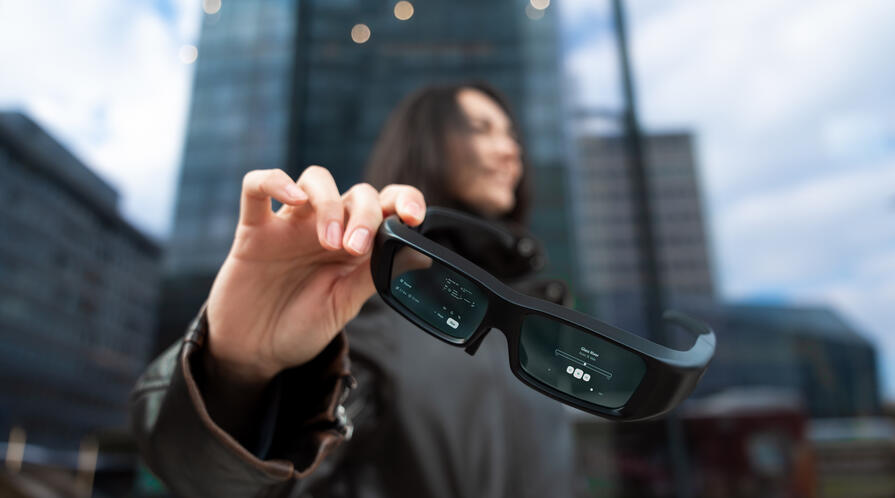 Close-up of smart glasses showing augmented reality interface while held by a woman in a modern urban environment.