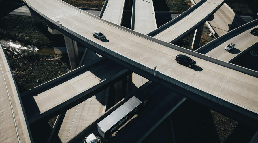 Aerial view of a multi-level highway interchange with sweeping curved overpasses crossing above one another. A few cars and a white semi-truck travel on the mostly empty concrete roads, while patches of vegetation and water are visible below the elevated ramps.
