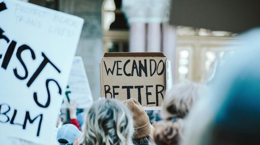 A crowd of people gathers at a protest, holding handmade signs. A cardboard sign in the center reads “WE CAN DO BETTER.”