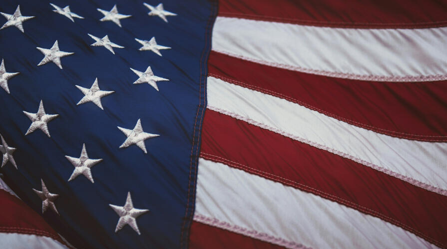Close-up photo of a rippling American flag, with embroidered white stars on a blue field on the left and red-and-white stripes waving on the right.