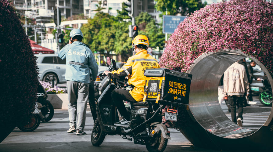 Two delivery workers, one on a scooter, wait outside a building in an urban setting in Shenzhen, China.