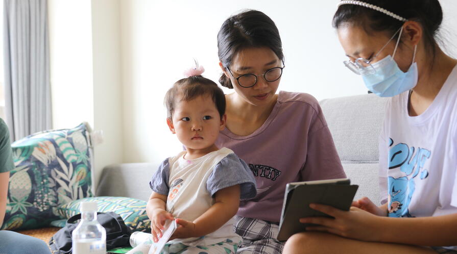 Migrant child sits on her mother’s lap, looking into the camera, as her mother reviews REAP’s survey forms with an enumerator. 