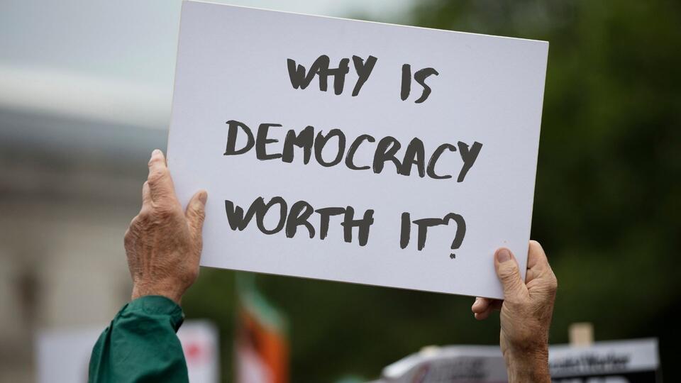 Hands holding a sign reading "why is democracy worth it?"