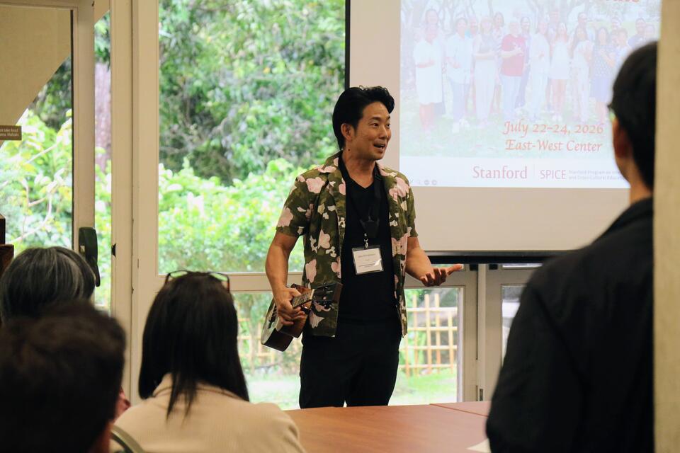 Man holding an 'ukulele and speaking to an audience